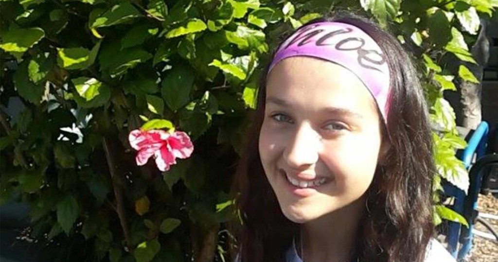 A girl with purple headband and pink flower in front of bush.
